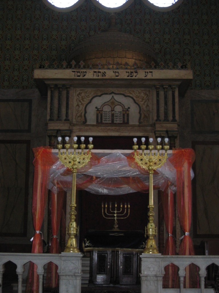 Colour image of synagogue interior featuring canopy made of white and orange gauze in front of wooden doors featuring the seven-branched candelabra. 