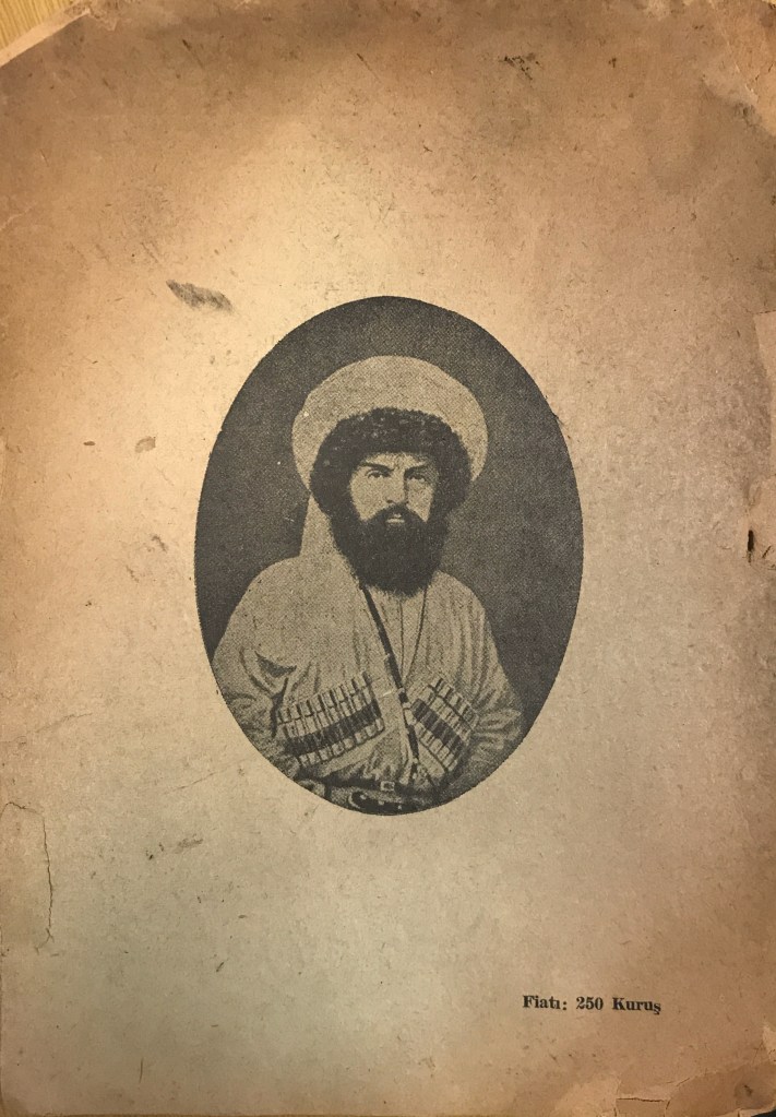 A black and white photograph of a man in traditional North Caucasian dress, looking directly into the camera, with a large sheep skin hat and bandoliers. 