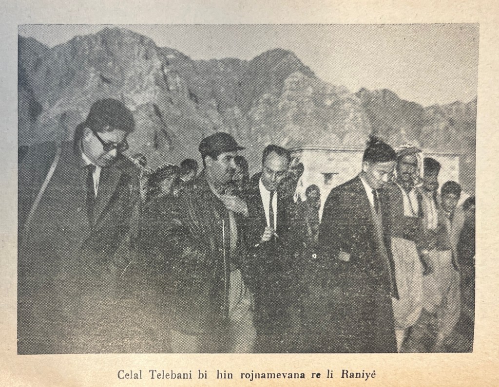 A group of men in European and Kurdish dress walking together towards something outside of the shot. Some are talking together. In the background mountains can be seen.