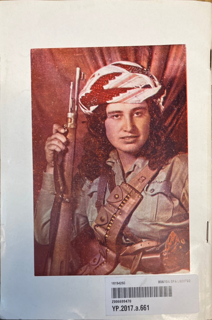 A colour photo of a woman in military fatigues and a Kurdish man's headdress with her curly red hair coming down from the bottom of the headgear. She is looking directly at the camera and pictured from the waist up brandishing a rifle. A carabiner is across her chest.
