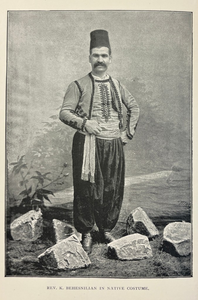 A black and white photo of a man in Armenian dress wearing a fez, a small waistcoat over a shirt, a sash around his waist and baggy trousers, with various broken stones around his feet