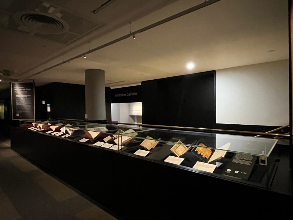 A darkened room with lit white walls to the right and four glass cases in the mid- and foreground with books in them 