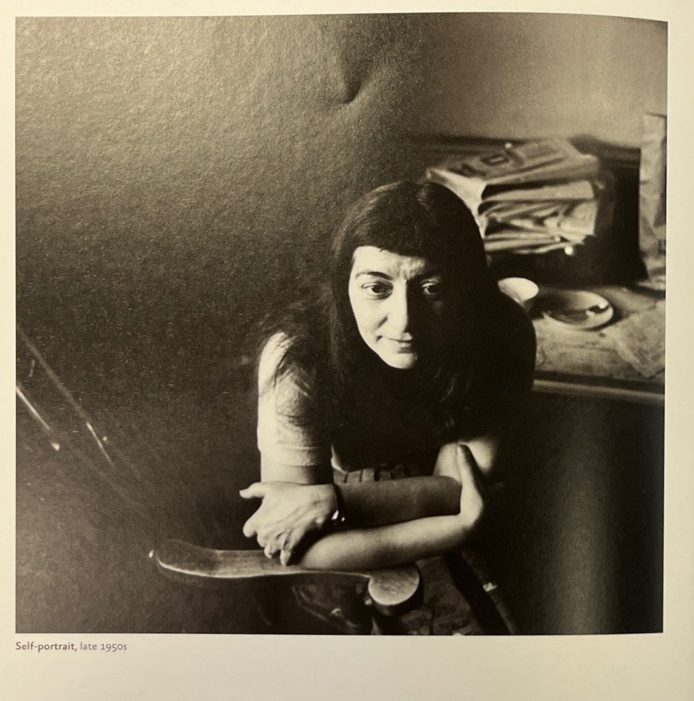 A black and white photo of a woman with dark long hair leaning on a chair arm with her arms crossed, looking just below the camera, in front of a padded bench with a dishes and stacked newspapers. 