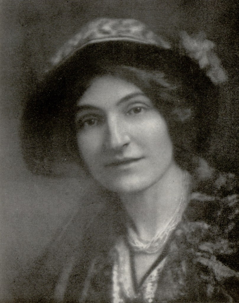 A black and white photograph of a woman around 40 looking into the camera. She is a wearing a large hat with a flower on its left side and her hair is drawn up under the hat. The frame covers from her chest up, showing a necklace, collar of her coat and décolleté